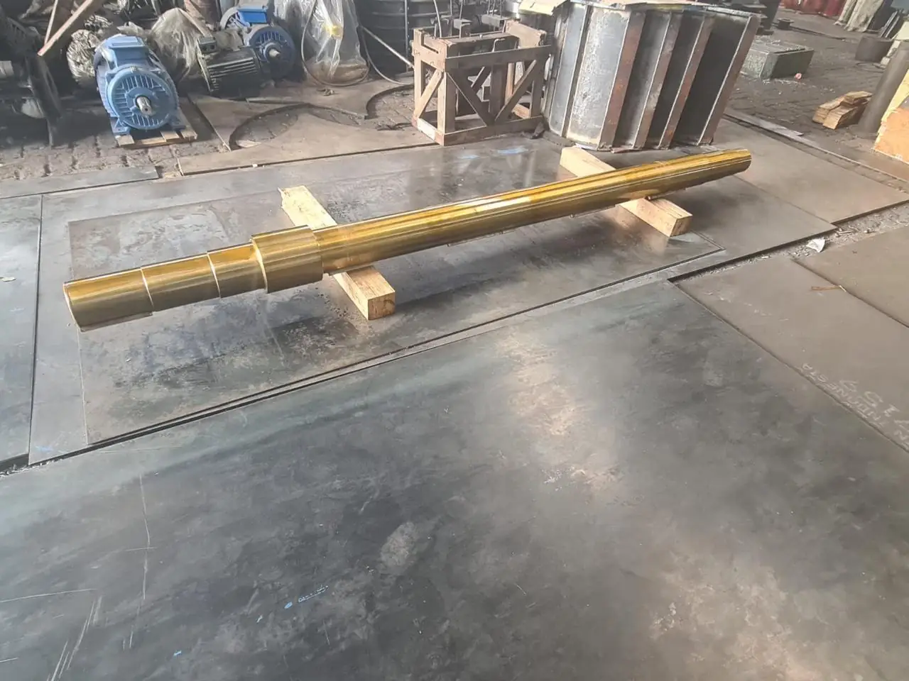 Long machined stepped shaft with a gold-colored finish resting on wooden blocks in a workshop